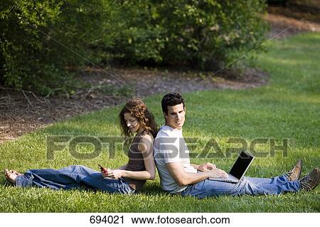 A young couple sitting back to back in grass using wireless devices View Large Photo Image Stock Image - A young couple sitting back to back in grass using wireless devices. Fotosearch