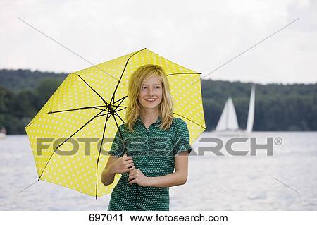 Stock Image - A young girl standing by a lake under an umbrella, overcast. Fotosearch