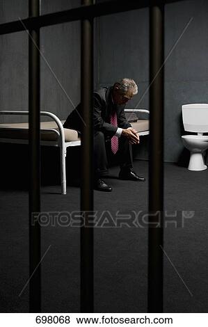 Stock Photo - A businessman sitting on a bed in a prison cell. Fotosearch