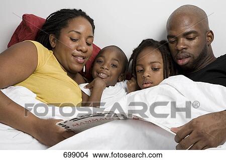 Picture - A family of four lying in bed reading a children's book. Fotosearch