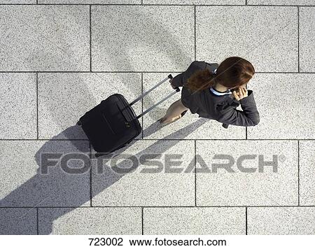 Stock Image - A businesswoman pulling a suitcase and talking on her mobile phone. Fotosearch