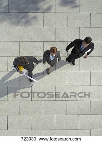 Stock Image - Three business people walking with a hard hat and blueprint. Fotosearch