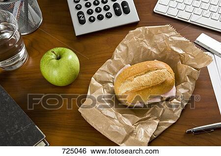 Stock Photograph - An office desk with office supplies and a sandwich and an apple on it. Fotosearch