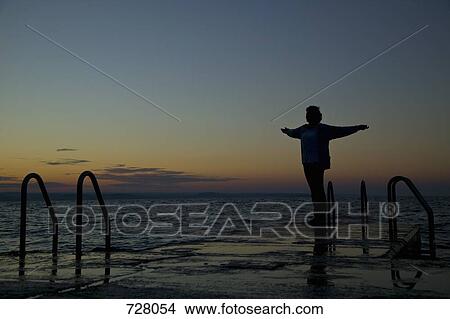 Picture - A woman standing at the edge of a jetty with her arms raised. Fotosearch