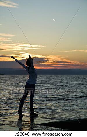 Stock Photograph - A woman standing at the edge of a jetty with her arms raised. Fotosearch