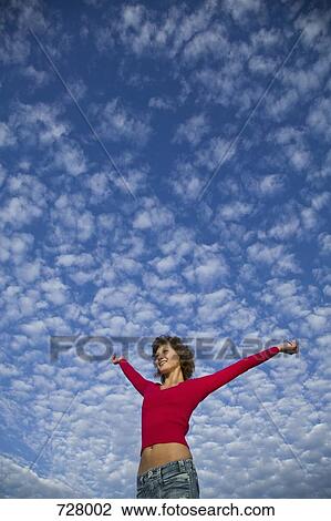 Stock Image - A woman with her arms raised against a cloudy sky, low angle view. Fotosearch