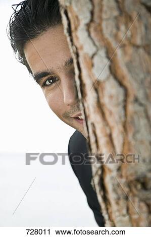 Stock Image - A young man peeking from behind a tree. Fotosearch