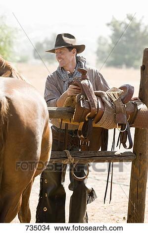 Picture - A rancher preparing a horse saddle on a fence. Fotosearch