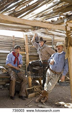 Stock Image - Three cowboys standing inside a wooden shed. Fotosearch