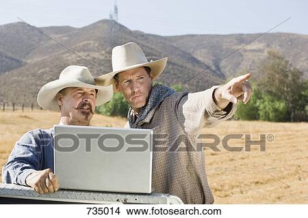 Picture - Two cowboys using a laptop and pointing. Fotosearch