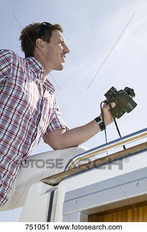 Stock Image - Side view of a man holding binoculars on a yacht. Fotosearch