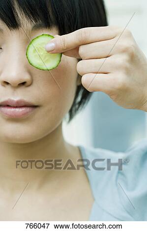 Stock Photo - A woman holding a slice of cucumber in front of her eye. Fotosearch