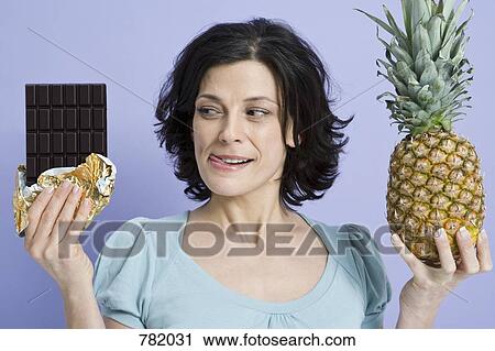 Stock Image - A woman deciding between a pineapple and a bar of chocolate. Fotosearch
