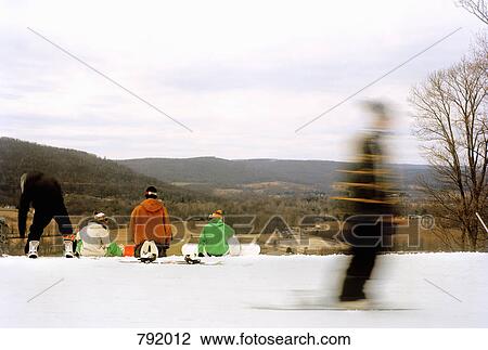 Stock Image - Rear view of snowboarders sitting at the edge of a ski slope. Fotosearch