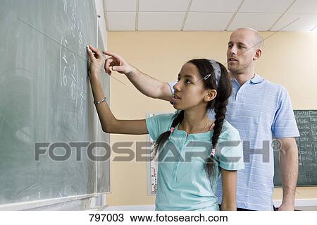 Stock Image - A girl writing on a blackboard with a teacher standing beside. Fotosearch
