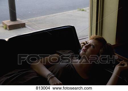 Picture - A young woman lying down on a sofa next to a window. Fotosearch