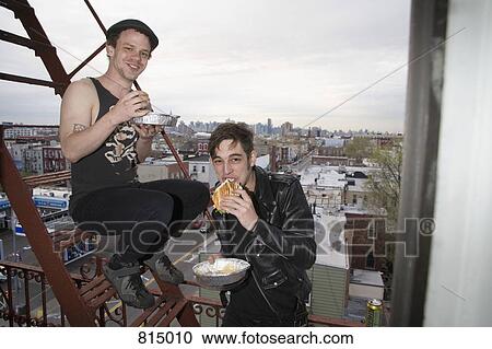 Stock Image - Two young men eating take out food on a fire escape. Fotosearch