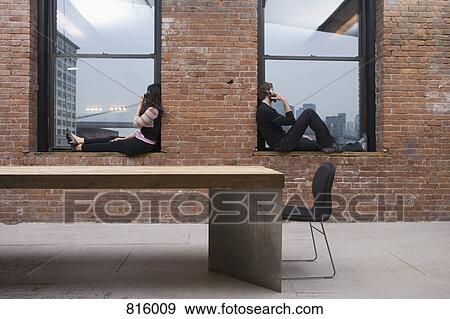Stock Photo - A man and woman sitting on window sills and talking on mobile phones. Fotosearch