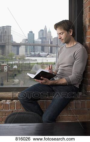 Stock Photograph - A man sitting on a window sill and looking through his person organizer. Fotosearch