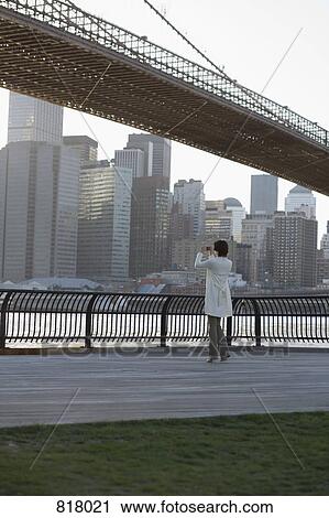 Stock Image - A woman taking a photograph near the Brooklyn Bridge, New York City. Fotosearch