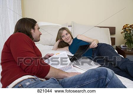 A young couple looking at magazines whilst lying on a bed View Large Photo Image Picture - A young couple looking at magazines whilst lying on a bed. Fotosearch