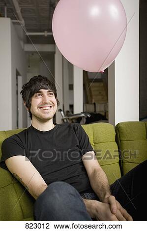 Stock Image - A pink balloon floating above a man sitting on a sofa. Fotosearch