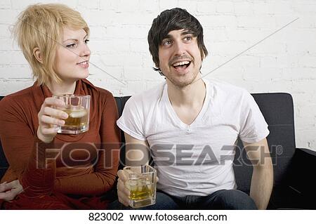 Stock Image - A young couple sitting on a sofa holding drinks. Fotosearch