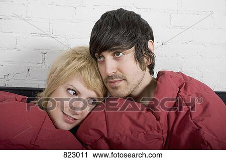 Stock Image - A young couple sitting on a sofa together under a blanket. Fotosearch
