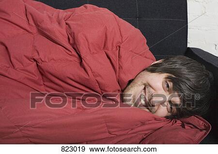 A young man lying under a blanket on a sofa View Large Photo Image Stock Photo - A young man lying under a blanket on a sofa. Fotosearch