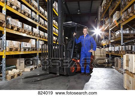 Stock Photo - A manual worker standing next to a forklift in a metal parts warehouse. Fotosearch