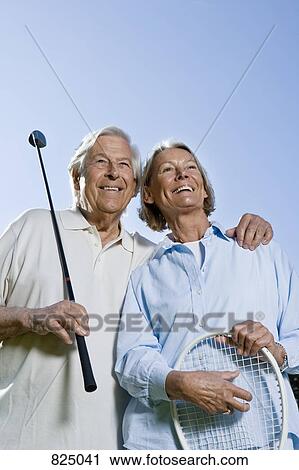 Stock Image - A senior couple holding a golf club and a tennis racket. Fotosearch