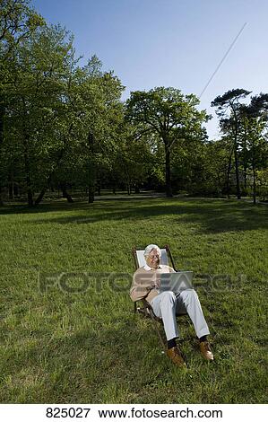 Stock Photo - A senior man sitting in a lounge chair using a laptop, outdoors. Fotosearch