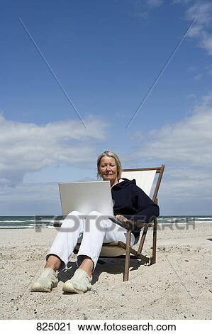 Stock Image - A senior woman sitting in a lounge chair at the beach using a laptop. Fotosearch