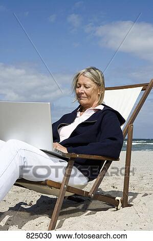 Stock Image - A senior woman sitting in a lounge chair at the beach using a laptop. Fotosearch