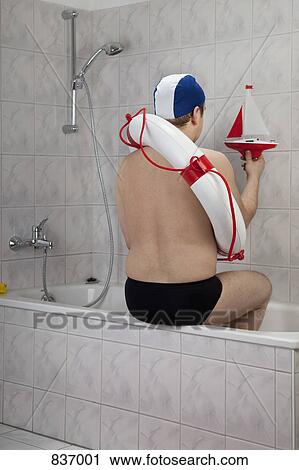 Stock Image - A man wearing racing briefs and a life preserver sitting on the edge of a bathtub, indoors. Fotosearch
