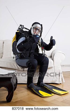Stock Photo - A scuba diver sitting on a couch in a living room giving a thumbs up sign. Fotosearch