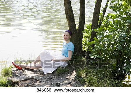 A young woman leaning against a tree next to a lake View Large Photo Image Stock Photograph - A young woman leaning against a tree next to a lake. Fotosearch