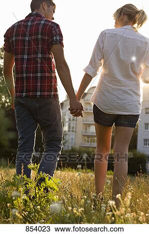 Stock Image - A young couple holding hands walking through a park, rear view. Fotosearch