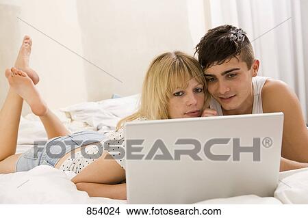A young couple lying on a bed using a laptop View Large Photo Image Picture - A young couple lying on a bed using a laptop. Fotosearch