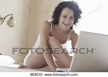 Stock Photo - A woman sitting on her bed and using her laptop. Fotosearch