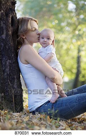 Stock Image - A mother sitting against a tree kissing her baby. Fotosearch