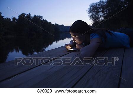 Stock Photo - A woman using an electronic organizer while lying on a jetty, dusk. Fotosearch
