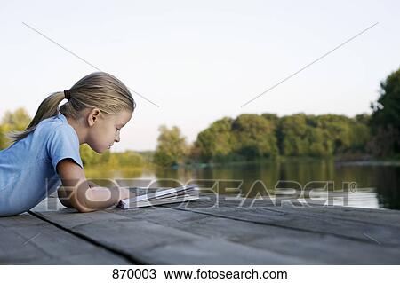 Stock Image - A young girl reading a book on a wooden jetty. Fotosearch