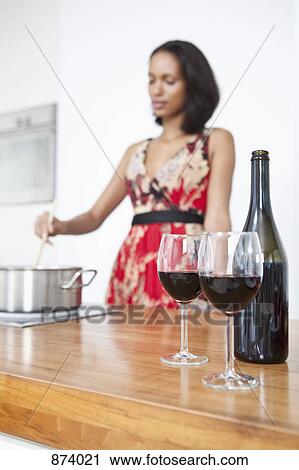 Stock Image - Two glasses of red wine and a wine bottle with a woman stirring a pot in the background. Fotosearch