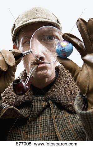 Stock Photography - A man dressed up as Sherlock Holmes looking at a tiny globe through a magnifying glass. Fotosearch