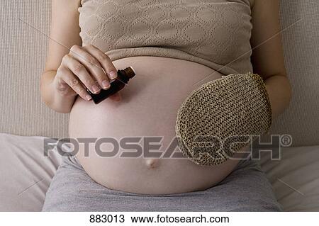 Stock Image - A pregnant woman applying body oil to her stomach, midsection. Fotosearch