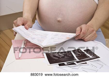 Stock Photo - A pregnant woman looking over medical paperwork and ultrasounds, midsection. Fotosearch