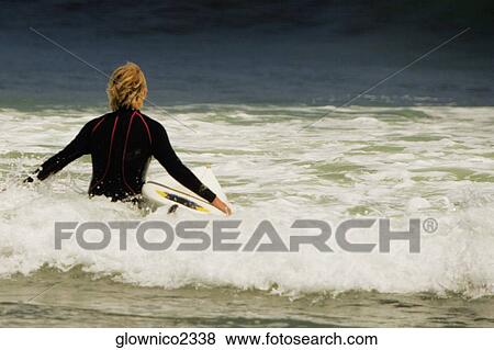 Stock Photo - Rear view of a man walking into the sea with a surfboard. Fotosearch