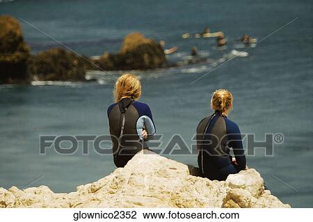 Stock Image - Rear view of two women sitting on a rock by the sea. Fotosearch