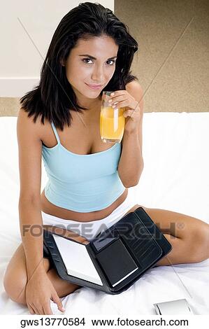 Portrait of a young woman sitting on a bed holding a glass of orange juice View Large Photo Image Picture - Portrait of a young woman sitting on a bed holding a glass of orange juice. Fotosearch